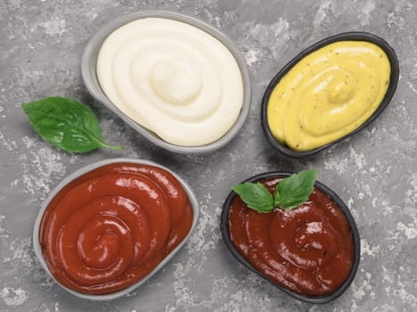 Top view of classic sauces set in trendy plates on gray concrete background. Sauces set - salsa, mustard, ketchup, mayonnaise, and basil for dressing. Flat lay.