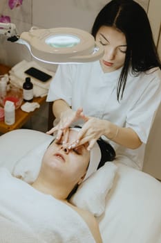 A young beautiful Caucasian brunette girl, a cosmetologist, gives a relaxing massage on the nose of an adult female client, who is lying with pleasure with her eyes closed on a massage table in a beauty salon, close-up side view.