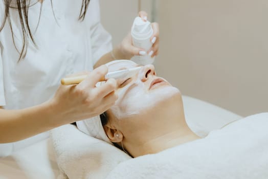 A young Caucasian girl, a cosmetologist, smears a white cream mask with a brush on the face of an adult female client in the nose area, who is lying with her eyes closed on a massage table in a beauty salon, side view close-up.