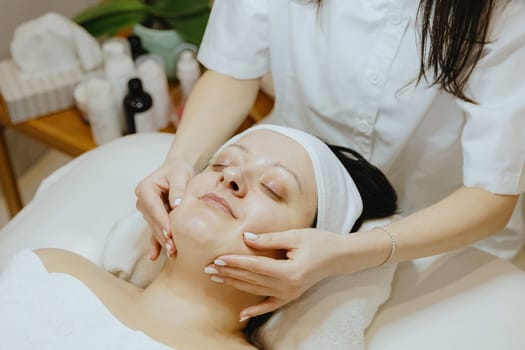 A young beautiful unrecognizable girl cosmetologist massages the face of an adult client in the chin area with her fingers, who lies contentedly with her eyes closed on a massage table in a beauty salon, close-up side view.
