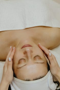 A young unrecognizable girl cosmetologist gently applies moisturizing cream with her fingers in the cheek area with stroking movements on the face of an adult client, who lies with her eyes closed on a massage table in a beauty salon, close-up side view.