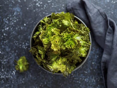 Green Kale Chips with salt in bowl. Homemade healthy snack for low carb, keto, low calorie diet. Dark blue background. Ready-to-eat kale chips, top view or flat lay