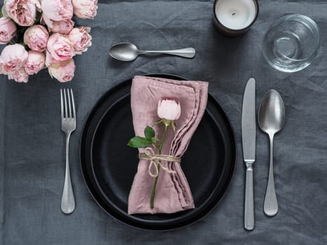 Beautiful table setting with candle on gray linen tablecloth. Festive table setting for wedding dinner with pink spray rose and pink napkin on plate. Holiday dinner with modern craft black plates