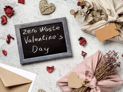 Zero waste Valentine's Day concept. Eco-friendly gift cloth wrapping in Furoshiki style and chalkboard with Zero Waste Valentine's Day letters on gray textured background. Top view or flat lay