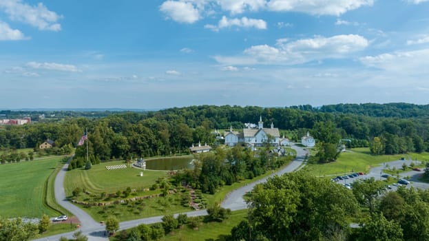 Expansive Aerial View Over A Religious Complex With Multiple White Buildings And Shining Golden Domes, A Pond With A Fountain, Set In A Verdant Landscape With A Large American Flag, With Fluffy Clouds