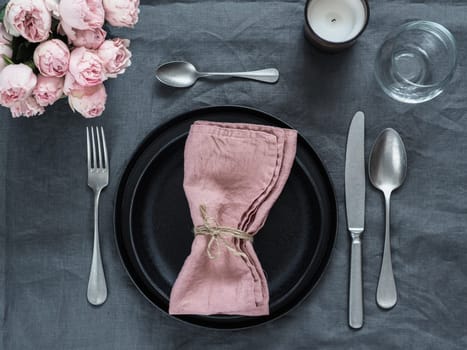 Beautiful table setting with pink spray roses and candle on gray linen tablecloth. Festive table setting for wedding dinner with pink napkin on plate. Holiday dinner with modern craft black plates