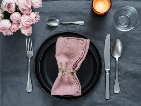 Beautiful table setting with pink spray roses and candle on gray linen tablecloth. Festive table setting for wedding dinner with pink napkin on plate. Holiday dinner with modern craft black plates