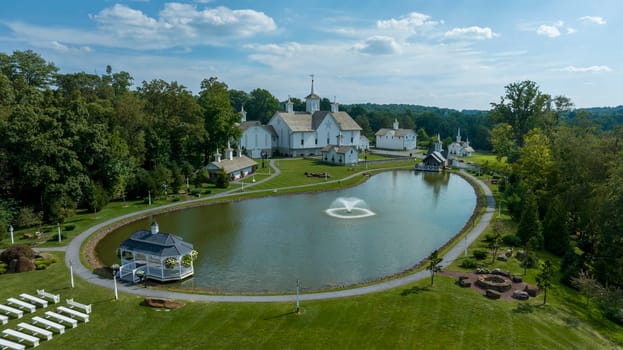 Elizabethtown, Pennsylvania, USA, August 11, 2023 - Aerial View Showcasing A Cluster Of Traditional White Orthodox Churches With Cross-Topped Domes, Arranged Around A Curved Pond With A Fountain, Amidst Green Trees And Neatly Arranged White Benches