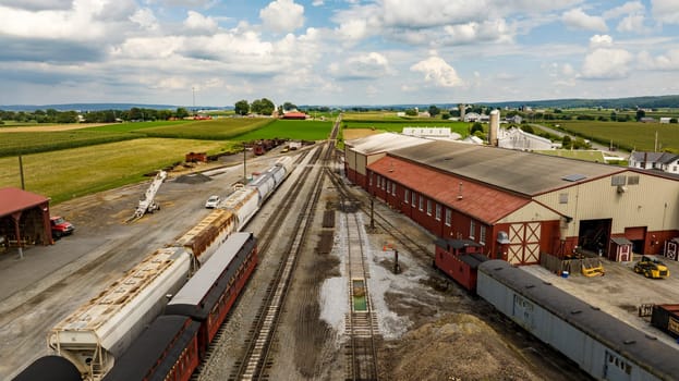 Strasburg, Pennsylvania, August 15, 2023 - An expansive view from above captures a rural rail yard with freight cars, surrounded by the lush, orderly farmlands stretching into the distance.