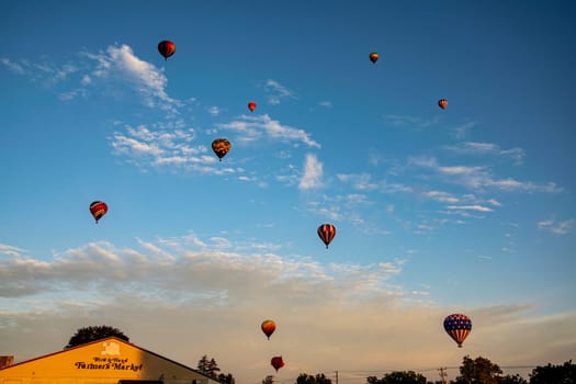 A fleet of vibrant hot air balloons takes to the sky over a local farmer's market at sunset, adding a touch of magic to the day's end.