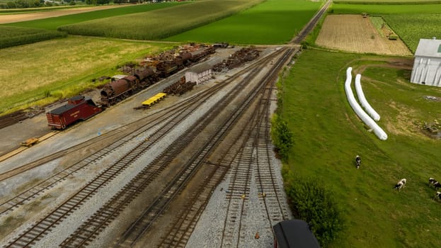 A rural train yard, scattered with vintage railcars, nestles among vast agricultural fields, showcasing a juxtaposition of industry and farming.