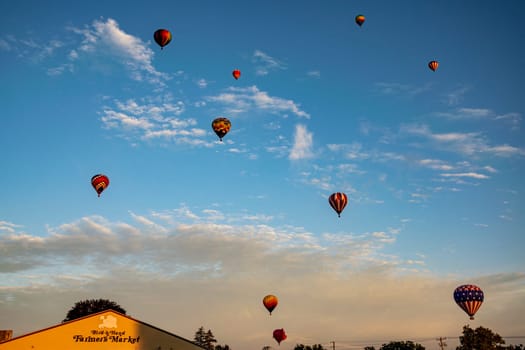 A fleet of vibrant hot air balloons takes to the sky over a local farmer's market at sunset, adding a touch of magic to the day's end.