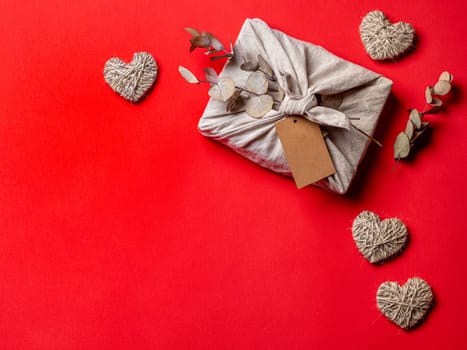 Zero waste, Valentine's Day eco-friendly gift wrapping in Furoshiki style with dry eucalypt and empty craft label. Gift box in cloth wrapping, on red background. Copy space for text.Top view, flat lay