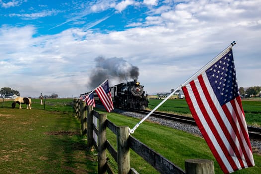 A vintage steam train chugs along the countryside, flanked by American flags on a wooden fence, evoking a sense of nostalgic Americana and historic travel.