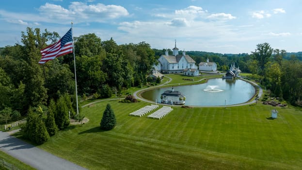 Elizabethtown, Pennsylvania, USA, August 11, 2023 - Aerial View Of A Grand White Building With A Grey Roof Surrounded By Lush Green Lawns, A Large Reflective Pond With Fountains, And A Majestic Flagpole Bearing The American Flag.