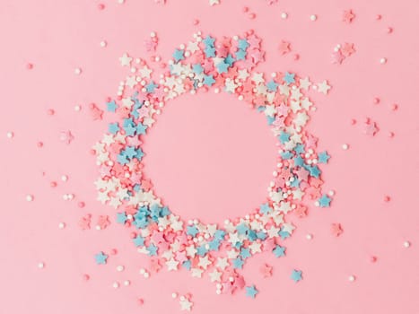 Festive border frame of colorful pastel sprinkles on pink background with copy space in center. Sugar sprinkle dots and stars in round shape, decoration for cake and bakery. Top view or flat lay