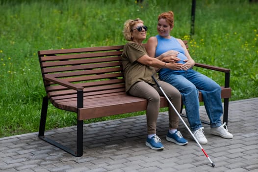 An elderly blind woman holds her pregnant daughter by the belly while sitting on a bench in the park