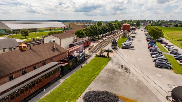 Strasburg, Pennsylvania, August 15, 2023 - A vintage steam train is poised for departure at a charming railway station, evoking a bygone era against a modern-day backdrop of cars and greenery.