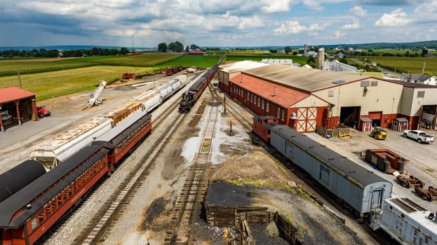 Strasburg, Pennsylvania, August 15, 2023 - Captured from above, a bustling railway junction services a mix of passenger and freight cars, framed by the lush farmland it supports.