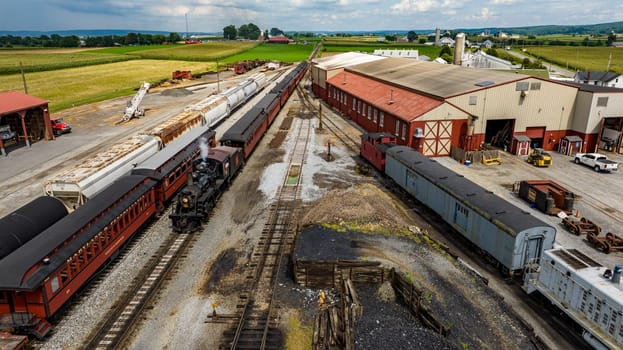 Strasburg, Pennsylvania, August 15, 2023 - Captured from above, a bustling railway junction services a mix of passenger and freight cars, framed by the lush farmland it supports.