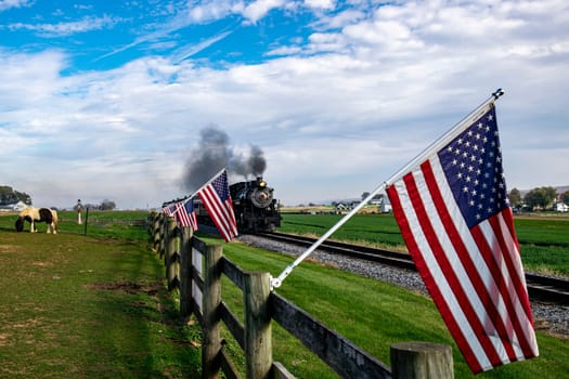 A vintage steam train chugs along the countryside, flanked by American flags on a wooden fence, evoking a sense of nostalgic Americana and historic travel.