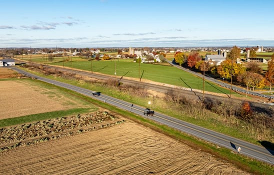 Late afternoon sun bathes a peaceful rural scene, featuring a horse-drawn buggy and vehicles on a country road, highlighting the blend of tradition and modernity.