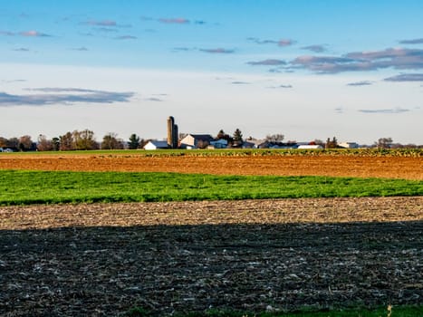 The cool dusk light washes over sprawling farmlands, highlighting the rich tapestry of tilled earth and burgeoning crops with a farmstead silhouette in the distance.