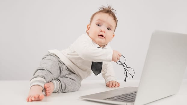 A cute child wearing a suit holds glasses and lies behind a laptop