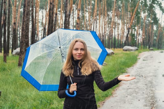 Young woman holding transparent blue umbrella outdoors in forest. Rainy weather day using umbrella. Woman with hand checking how long it will be raining. Forecast