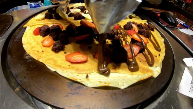 Delicious crepe being drizzled with chocolate sauce, topped with strawberries and brownie pieces