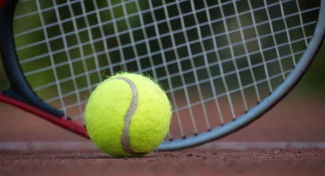 Close up of a yellow tennis ball and a tennis racket lying on a tennis hard court surface is depicted