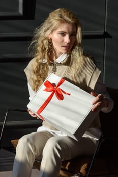 An artist is seated in a chair, showcasing a white giftbox with a red ribbon.