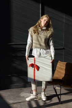 A woman holds a white gift box with a red ribbon. Model wearing stylish knitted vest, white shirt and classic trousers.