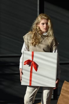 A woman holds a white gift box with a red ribbon. Model wearing stylish knitted vest, white shirt and classic trousers.