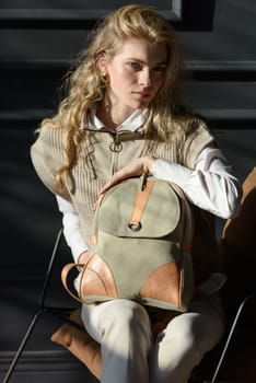 A woman holds leather backpack. Model wearing stylish knitted vest, white shirt and classic trousers.