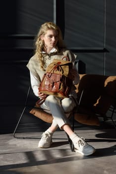 A woman holds leather backpack. Model wearing stylish knitted vest, white shirt and classic trousers.