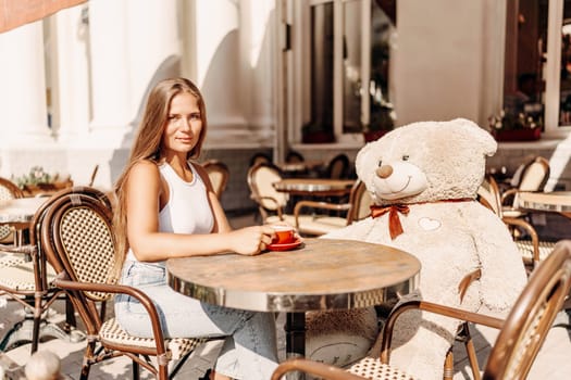 A woman sits cafe with a teddy bear next to her. The scene is set in a city with several chairs and tables around her. The woman is enjoying her time at the outdoor cafe