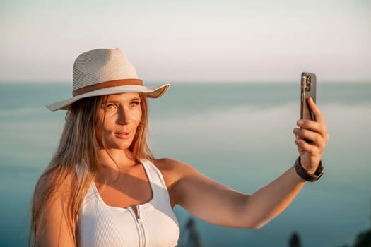 Selfie woman in hat, white tank top and shorts makes selfie shot mobile phone post photo social network outdoors on sea background beach people vacation lifestyle travel concept