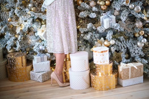 Women's legs. Cropped close-up photo of healthy beautiful elegant female legs in beige high-heeled shoes, a woman stands on the background of a Christmas tree with gifts.