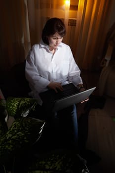 A girl in white shirt with a laptop in a dark room. A middle-aged woman works online remotely in the evening. The student is preparing for the exam at night. Blogger or freelancer in the chat