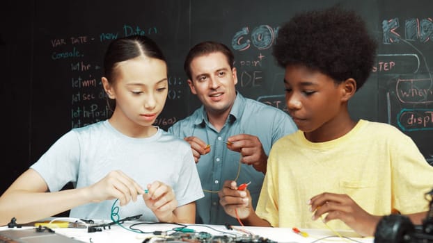 Caucasian teacher talking about electric tool while student fixing robotic model on table with electronic equipment placed. Diverse academic children learning about main board structure. Edification.