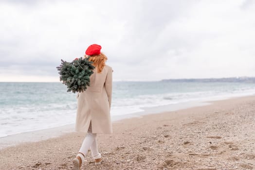 Redhead woman Christmas tree sea. Christmas portrait of a happy redhead woman walking along the beach and holding a Christmas tree on her shoulder. She is dressed in a light coat and a red beret