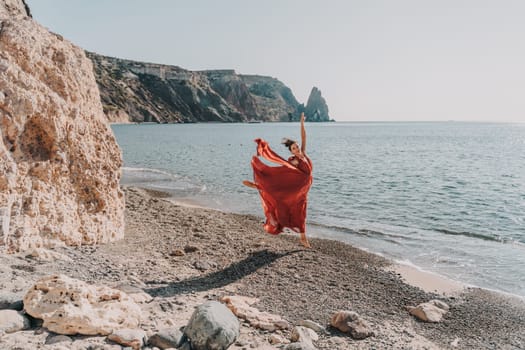 Woman red dress sea. Female dancer in a long red dress posing on a beach with rocks on sunny day. Girl on the nature on blue sky background