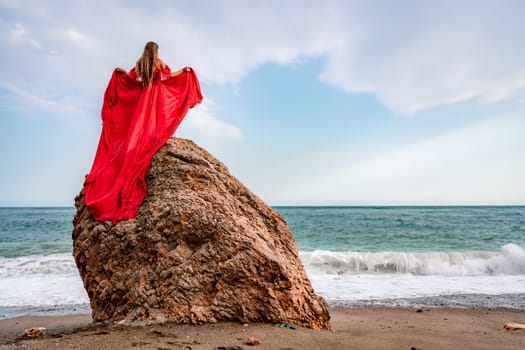 woman sea red dress. Woman with long hair on a sunny seashore in a red flowing dress, back view, silk fabric waving in the wind. Against the backdrop of the blue sky and mountains on the seashore