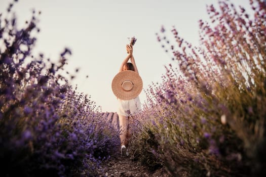 A woman is walking through a field of purple flowers with a straw hat on.