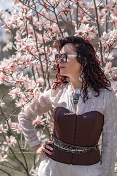 Magnolia park woman. Stylish woman in a hat stands near the magnolia bush in the park. Dressed in white corset pants and posing for the camera