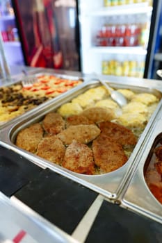 A bustling cafeteria serving line is filled with an array of dishes during a busy lunchtime. Trays of hot food are neatly arranged under the protective glass shield, with customers in the background selecting their meal choices. The variety suggests a diverse menu, catering to different tastes.