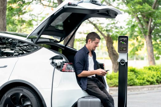 Man using smartphone online banking application to pay for electric car battery charging from EV charging station during vacation holiday road trip at national park or summer forest. Exalt