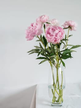 Beautiful pale pink peony bouquet in glass vase on table with white linen tablecloth. Vertical. Copy space for text. Toned filter