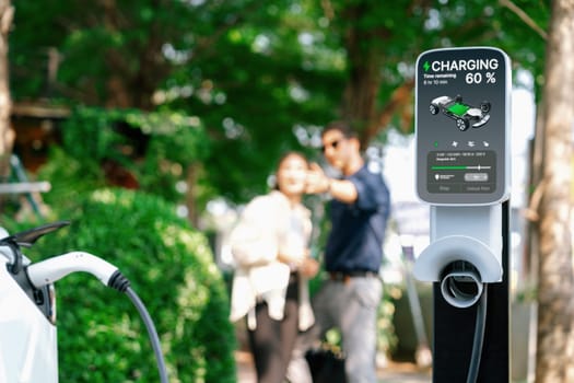 Young couple recharge electric car's battery from charging station in outdoor green city park in springtime. Rechargeable EV car for sustainable environmental friendly urban travel lifestyle.Expedient